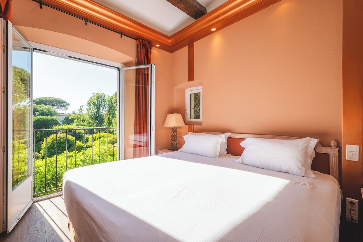 Habitación de hotel con una cama grande, una mesita de noche, una lámpara y vistas a zonas verdes a través de un ventanal corredero.