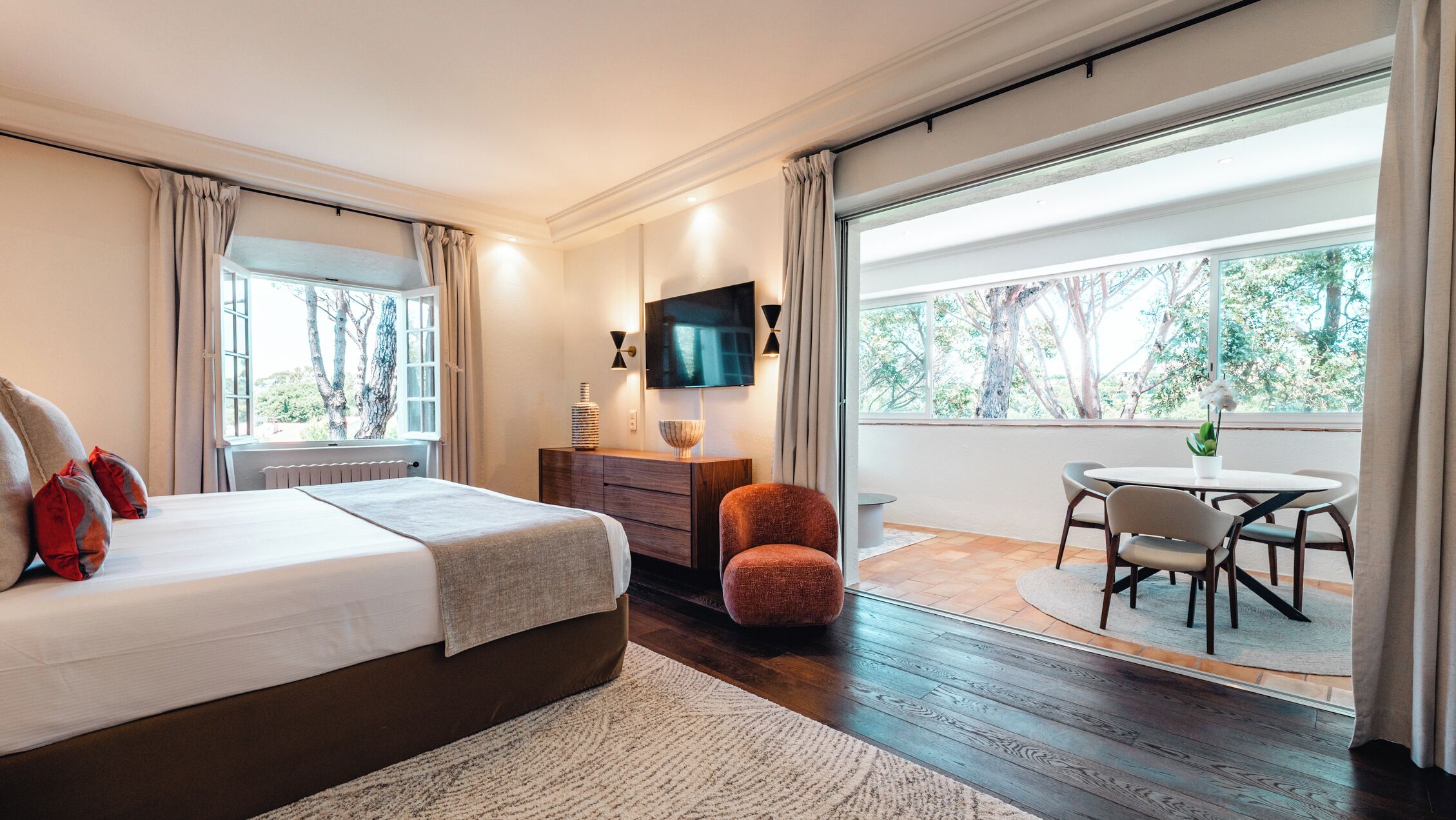 A modern bedroom with a bed, a TV, a small table, and a view of the outdoors.