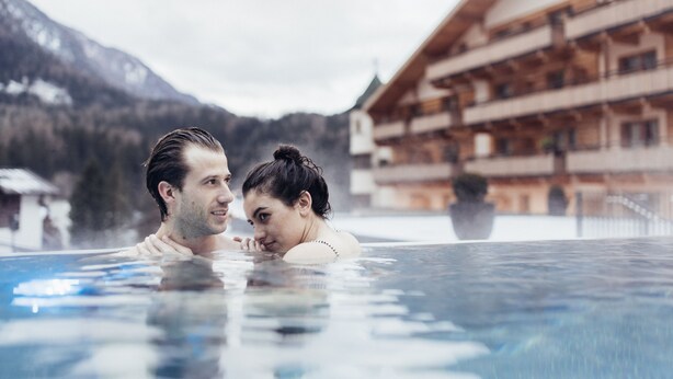 A couple in a hot tub with a mountain backdrop and a multi-story building. - Hotel Habicher Hof (Oetz)