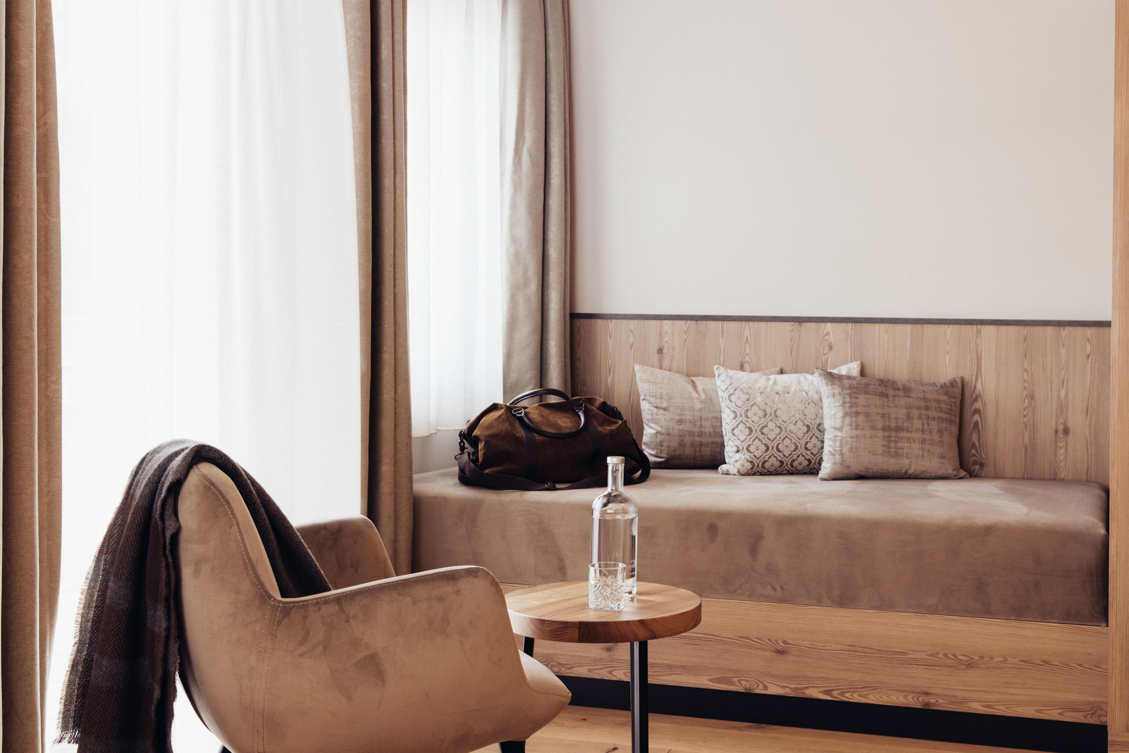 Een hotelkamer met een bank, een stoel, een klein rond tafeltje met een fles en een bruine tas op de bank.
