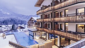 A multi-story hotel with a pool area, wooden balconies, and a view of snow-covered mountains. - Hotel Habicher Hof (Oetz)