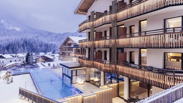 A multi-story hotel with a pool area, wooden balconies, and a view of snow-covered mountains. - Hotel Habicher Hof (Oetz)