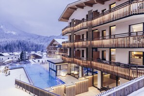 Ein mehrstöckiges Hotel mit Poolbereich, Holzbalkonen und Blick auf verschneite Berge.
