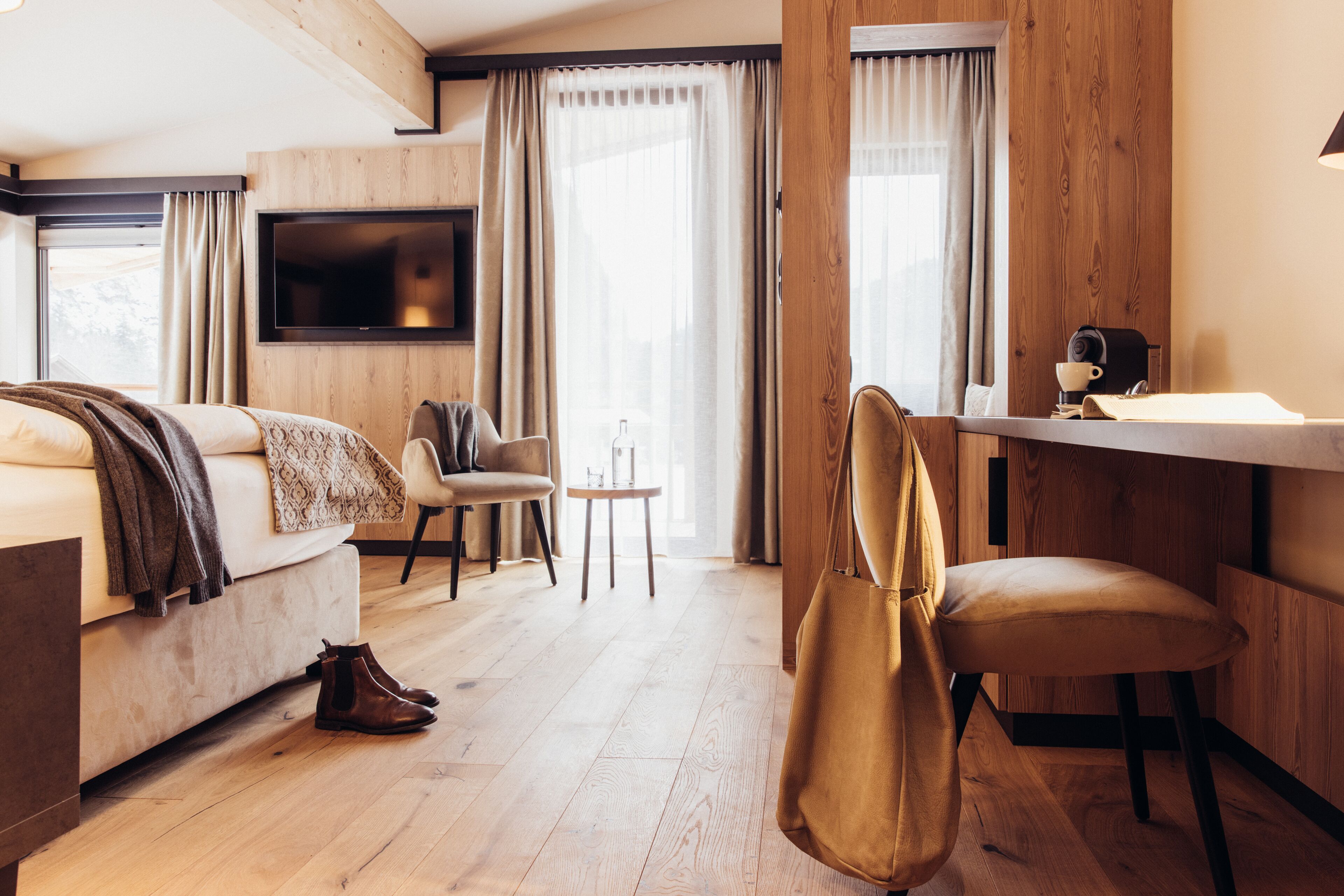A modern hotel room with a wooden floor, a sofa, a chair, a small table, a TV mounted on the wall, and a desk with a lamp.