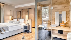 A modern hotel room with a large bed, bedside tables, and a glass partition leading to a sauna area with wooden benches and towels. - Hotel Habicher Hof (Oetz)