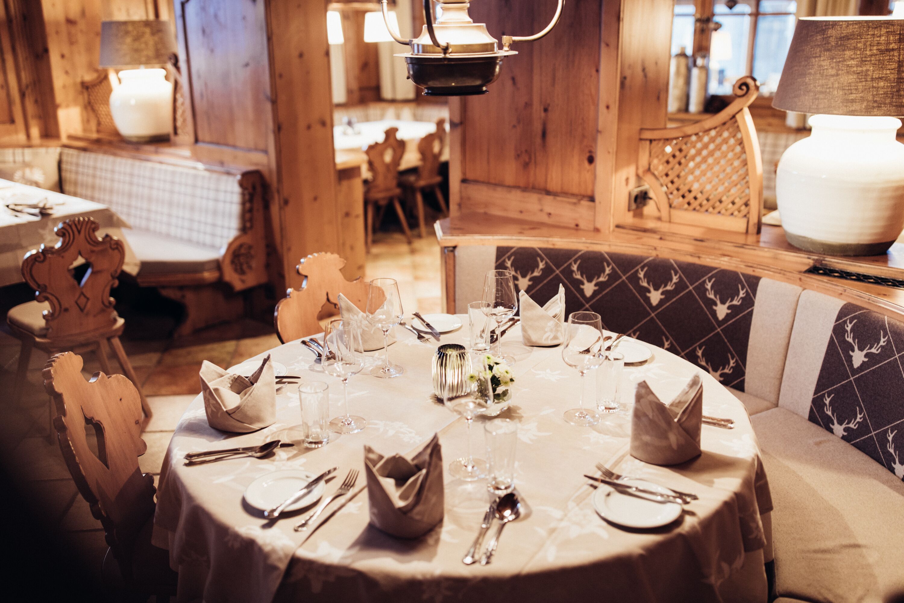 Mesa redonda de comedor con mantel blanco, cubiertos y copas, rodeada de sillas de madera con cojines estampados.
