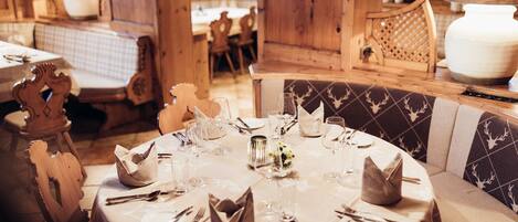 A round dining table set with white tablecloth, silverware, and glassware, surrounded by wooden chairs with a patterned cushion.