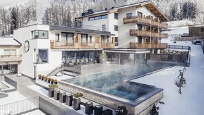 A modern hotel with a large outdoor pool, multiple balconies, and a view of snow-covered mountains. - Hotel Habicher Hof (Oetz)