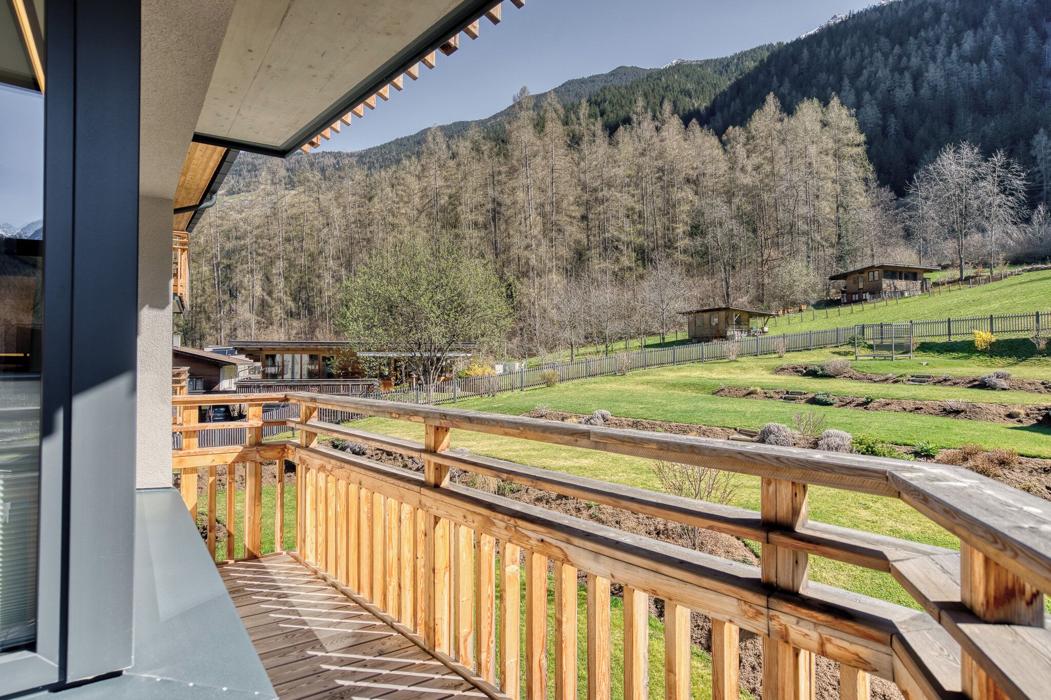 A wooden deck with a view of a mountainous landscape, a fenced garden, and a small building in the background.