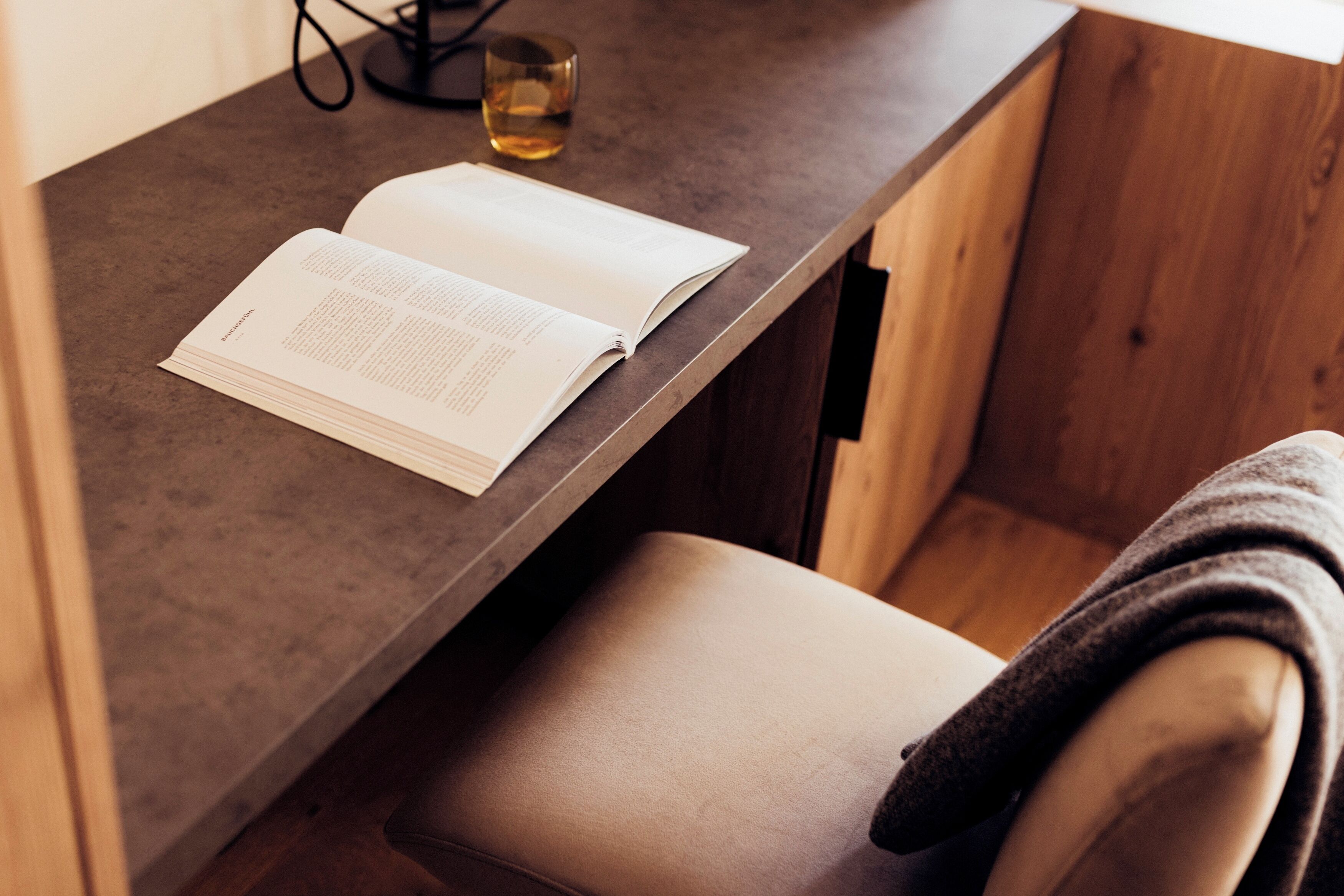 Um livro sobre uma mesa de madeira com um copo de líquido, uma cadeira e um armário de madeira.
