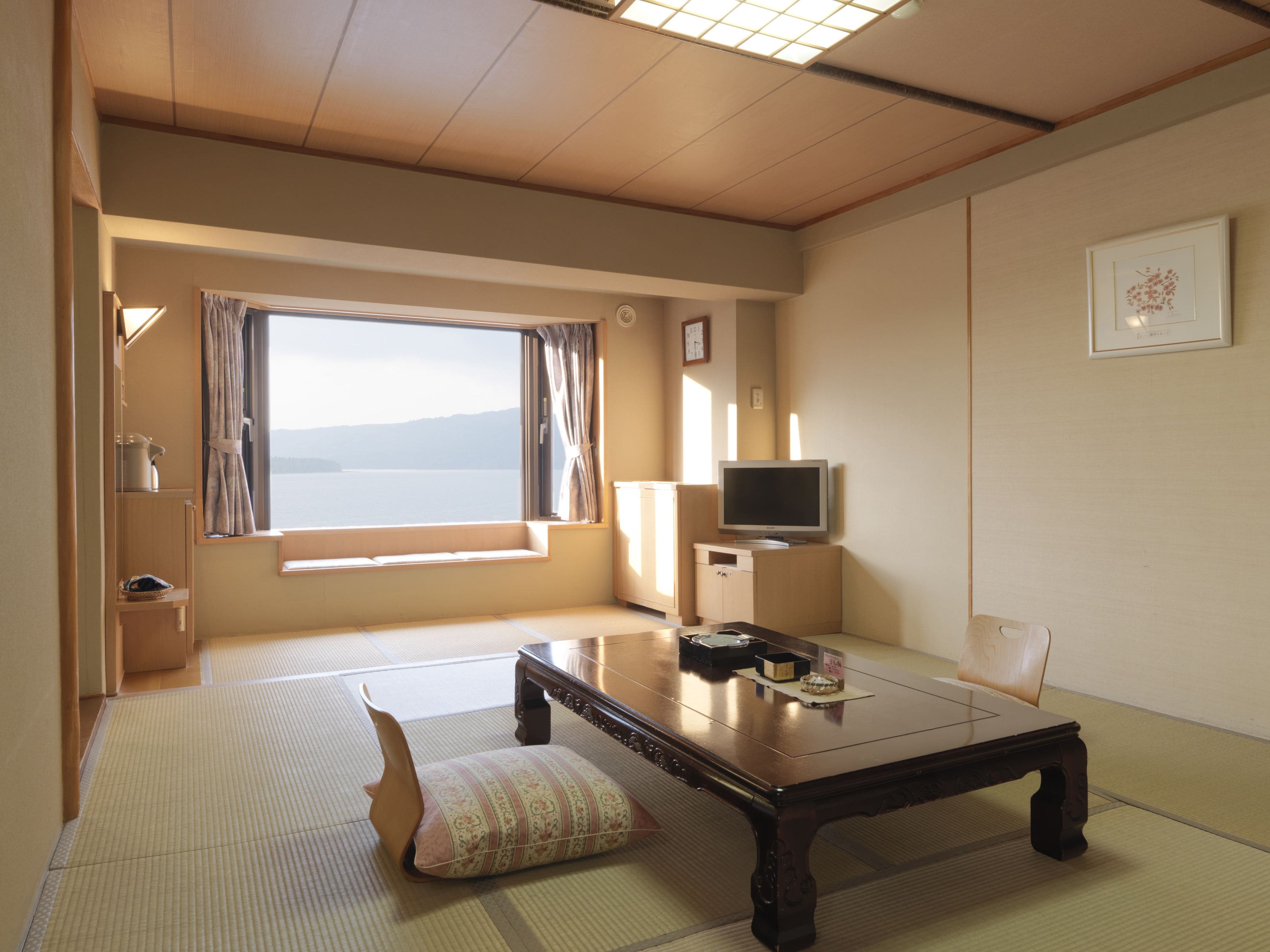 A room with tatami flooring, a low wooden table, a cushioned bench, a television on a cabinet, and a large window with a view of the sea.
