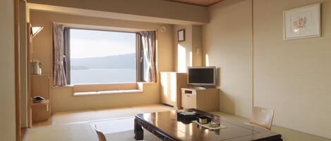 A room with tatami flooring, a low wooden table, a cushioned bench, a television on a cabinet, and a large window with a view of the sea.