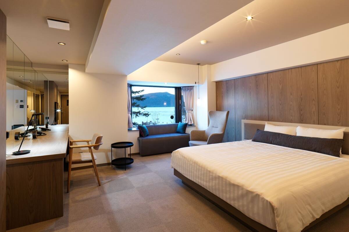 Une chambre d’hôtel moderne avec un grand lit, un bureau, une chaise et un canapé. Une fenêtre offre une vue sur un lac et des montagnes.