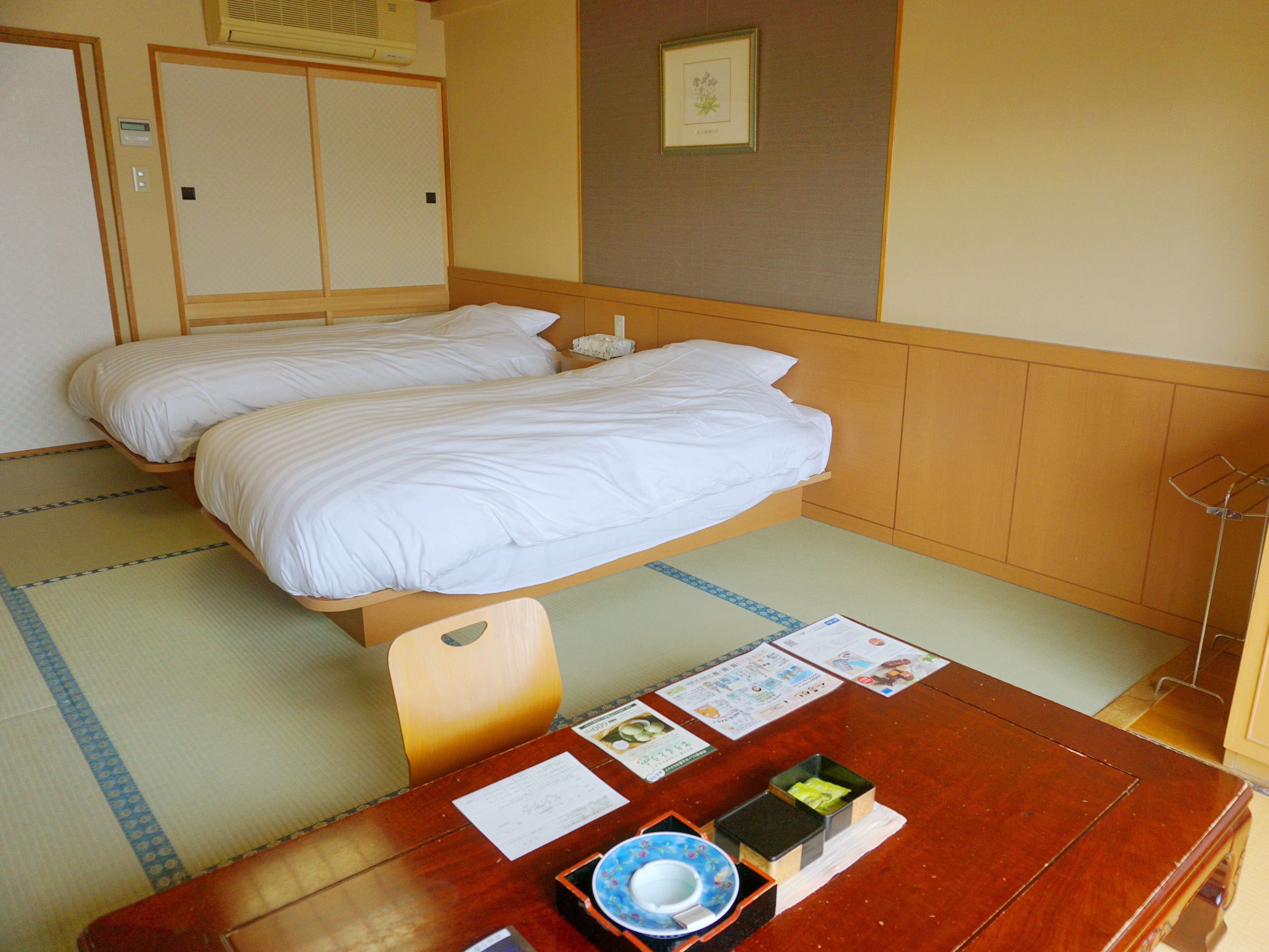 A hotel room with two beds, a wooden table with a bowl and papers, and a framed picture on the wall.