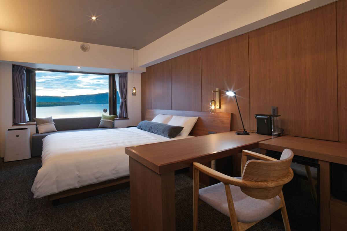 Une chambre d’hôtel moderne avec un grand lit, un bureau et une chaise. La fenêtre offre une vue sur un lac.