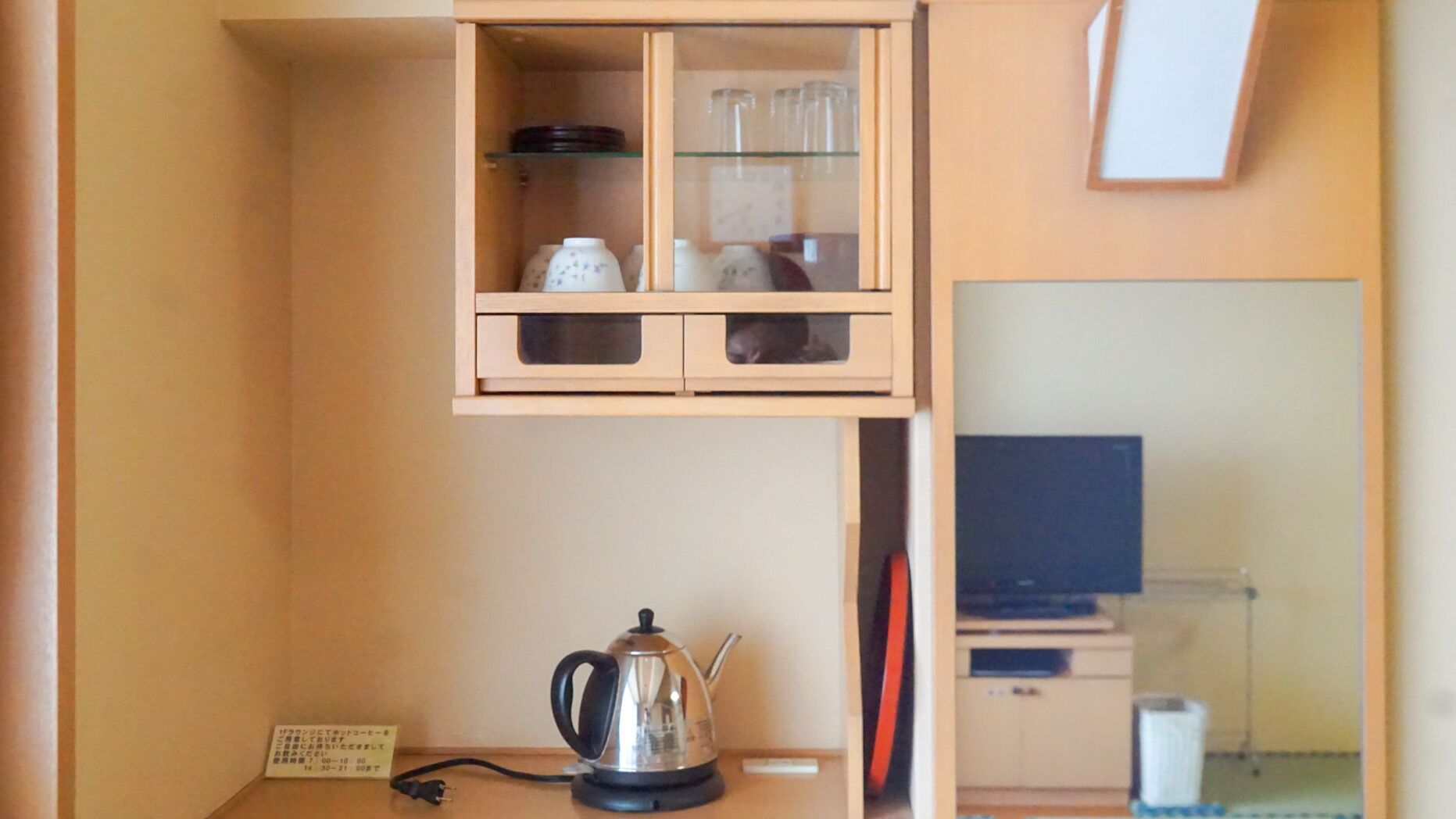 Un espace cuisine compact avec une bouilloire sur le comptoir, un petit meuble avec des portes en verre contenant des ustensiles de cuisine, et un téléviseur posé sur un meuble au fond de la pièce.