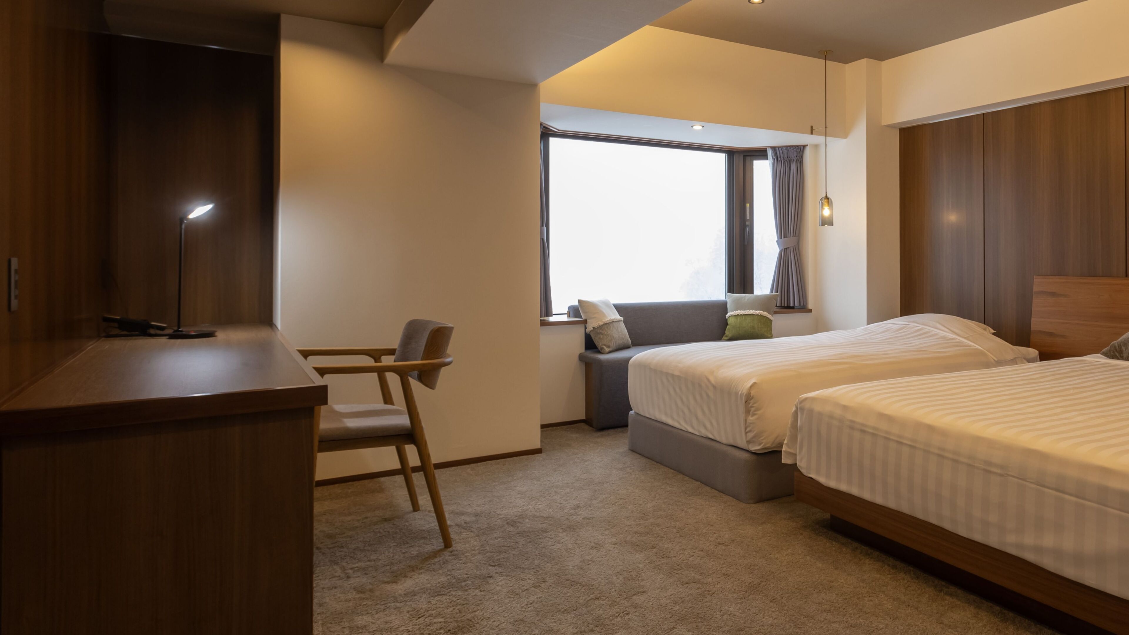 Ein Hotelzimmer mit Schreibtisch, zwei Betten, einem Fenster mit Vorhängen und Blick zum Himmel.