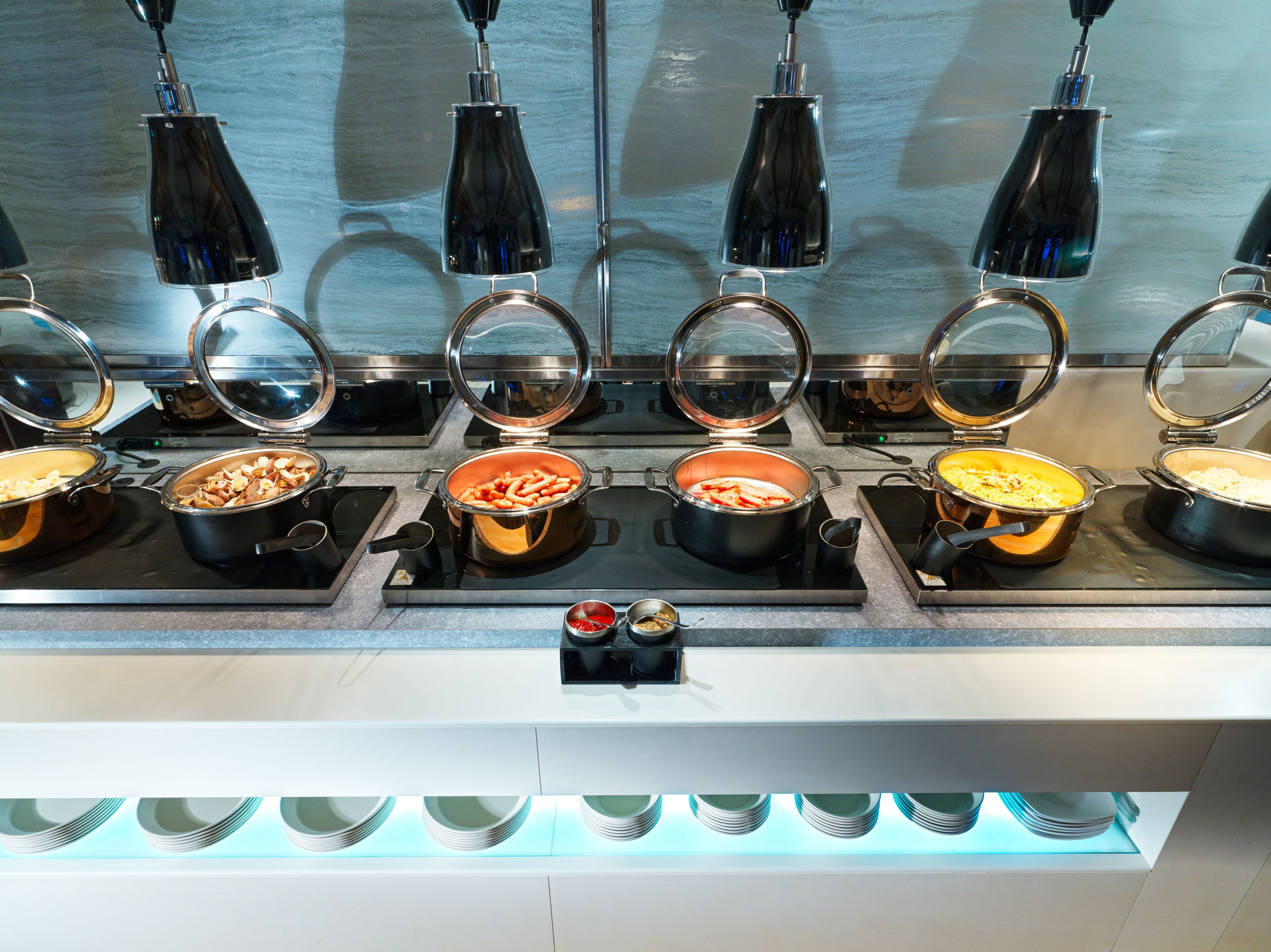 A buffet setup with multiple hot dishes, a central condiment station, and overhead lighting.