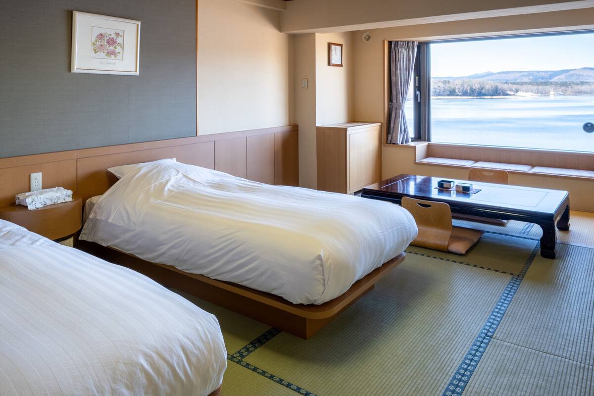Une chambre d’hôtel avec deux lits, une petite table, une fenêtre donnant sur un lac et une horloge au mur.