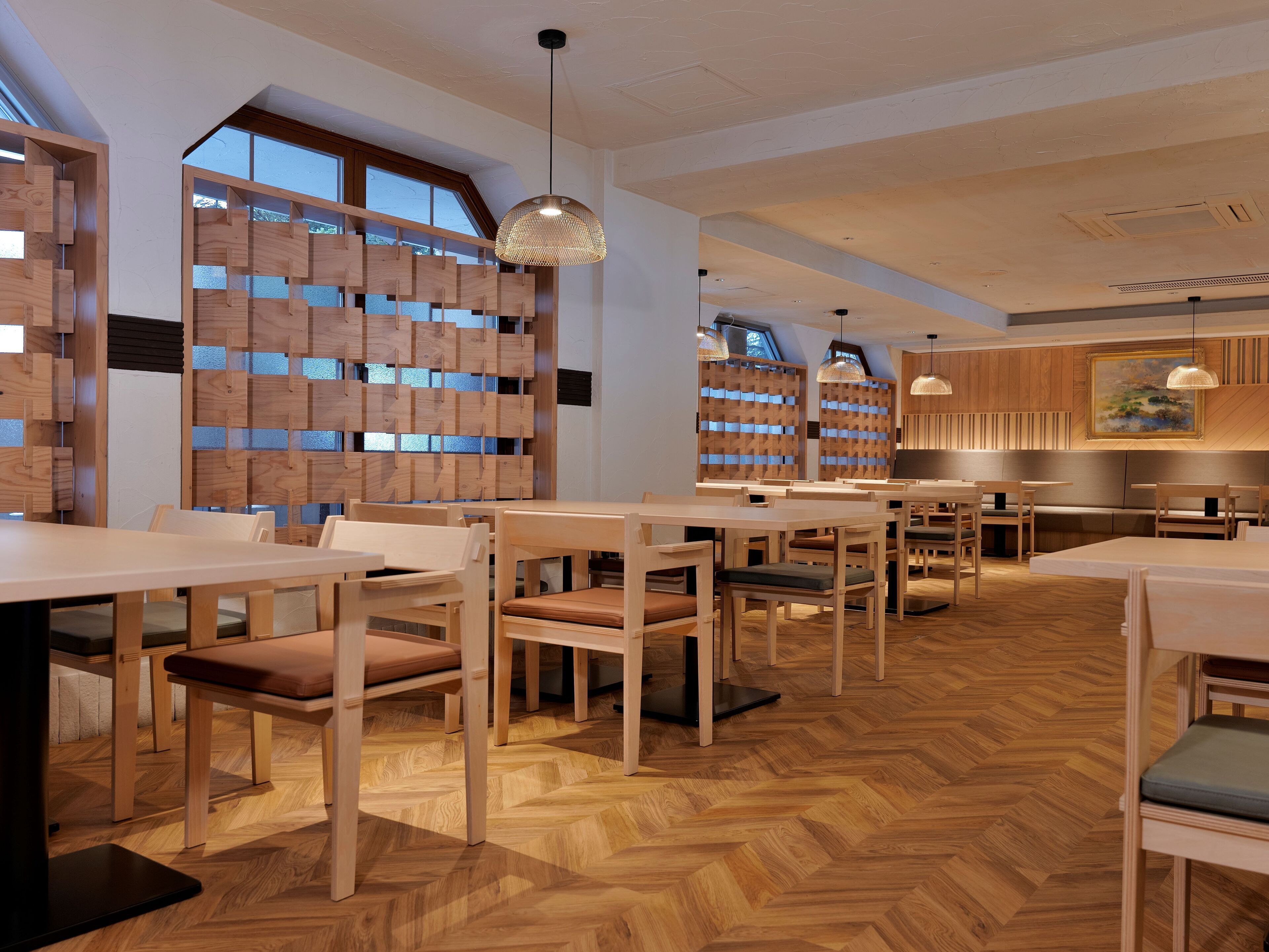 A modern restaurant interior with wooden furniture, herringbone-patterned flooring, and large windows allowing natural light.