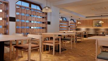 A modern restaurant interior with wooden furniture, herringbone-patterned flooring, and large windows allowing natural light.