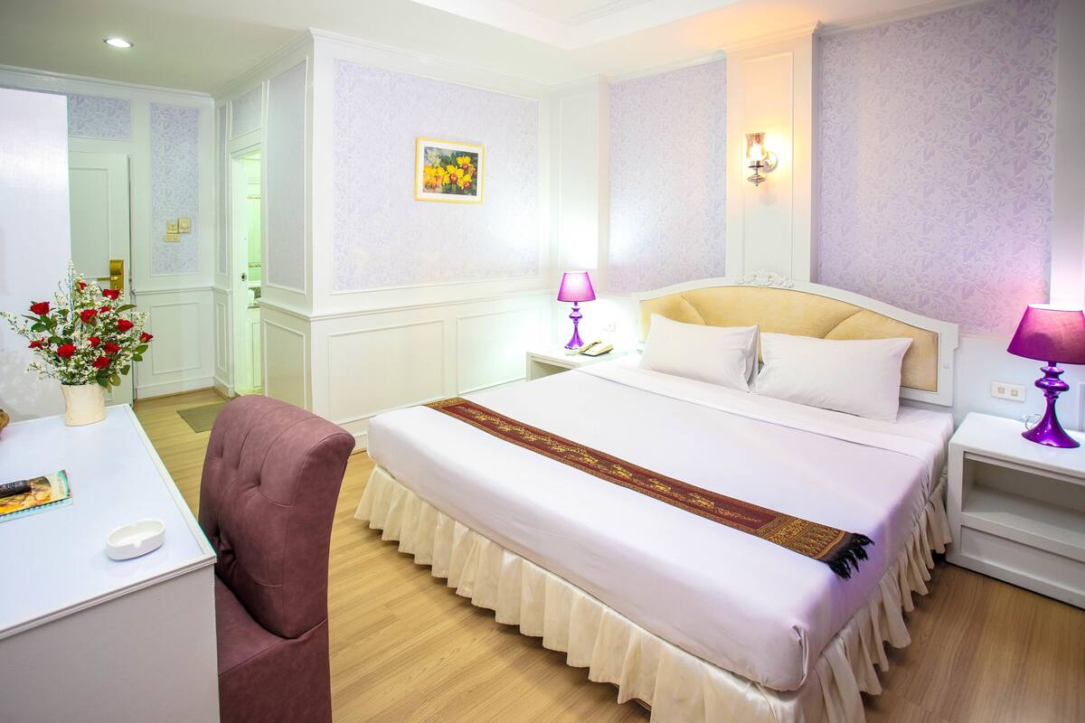 A hotel room with a large bed, a desk with a chair, a bedside table with a lamp, and a vase with flowers.