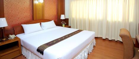 Quarto de hotel com uma cama grande, cabeceira de madeira, mesa de cabeceira com abajur e uma cadeira.