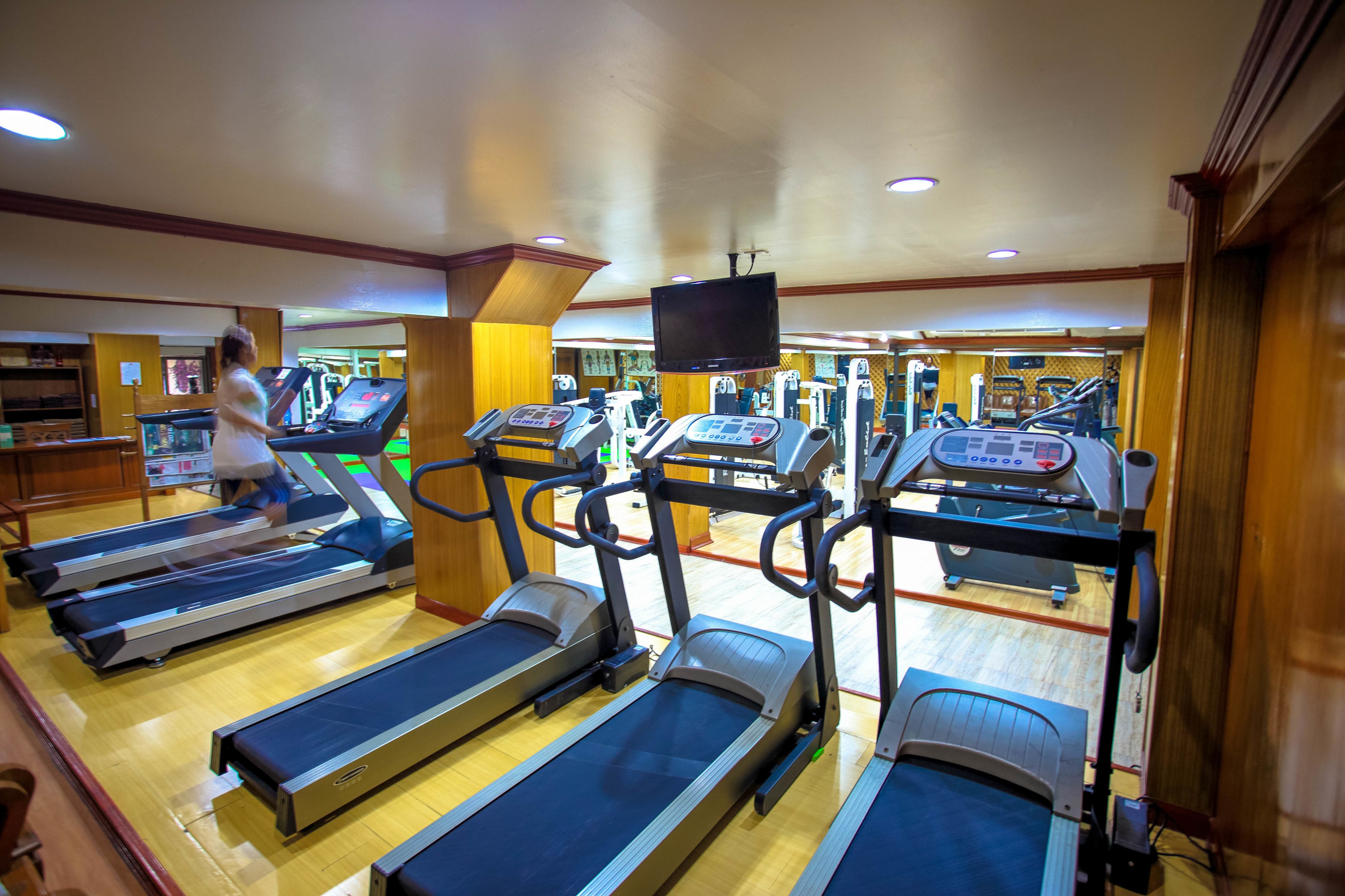 Een fitnessruimte met meerdere loopbanden, een aan het plafond gemonteerde televisie en houten lambrisering.