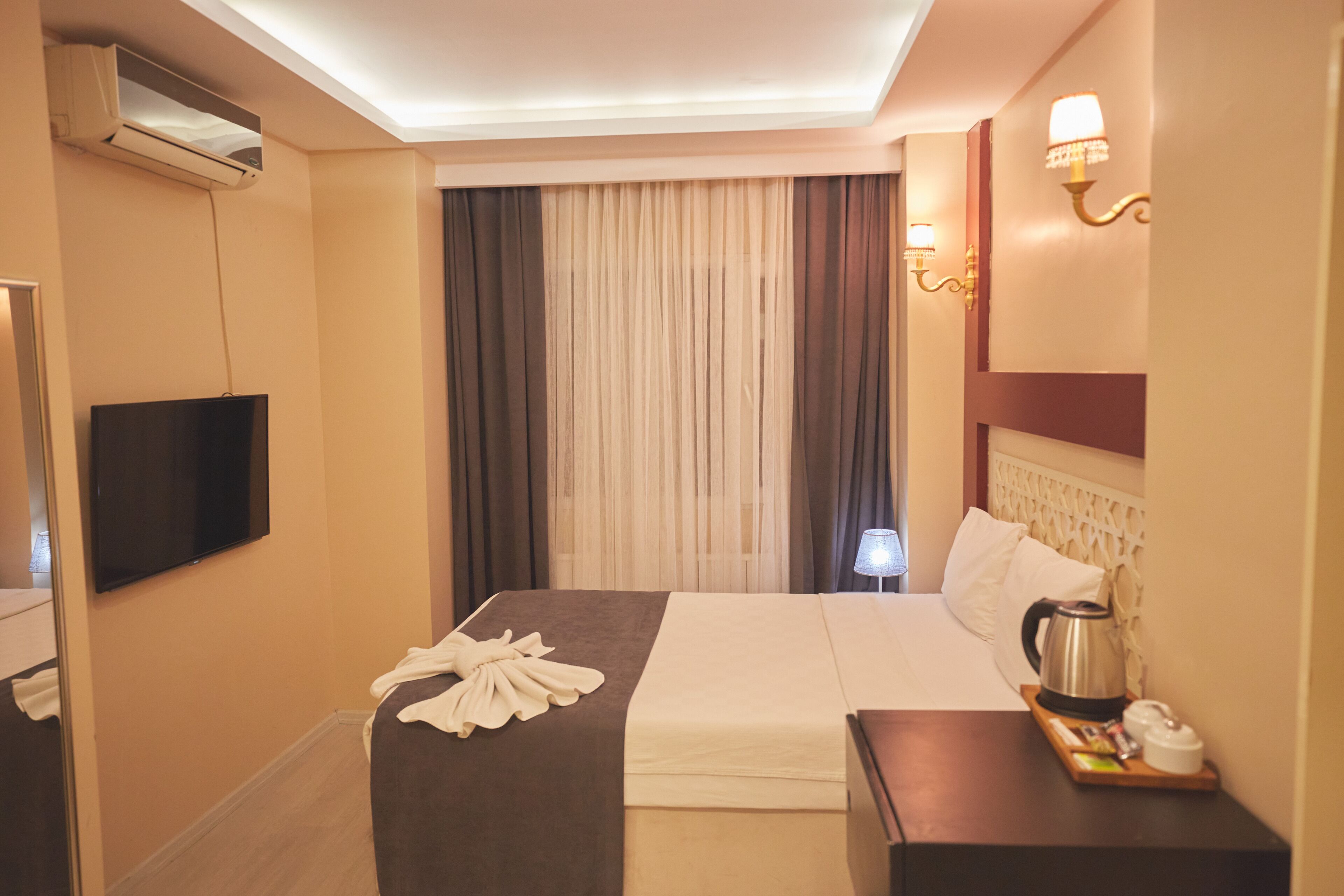 A hotel room with a bed, a flat-screen TV, an air conditioner, a bedside table with a kettle, and a lamp.