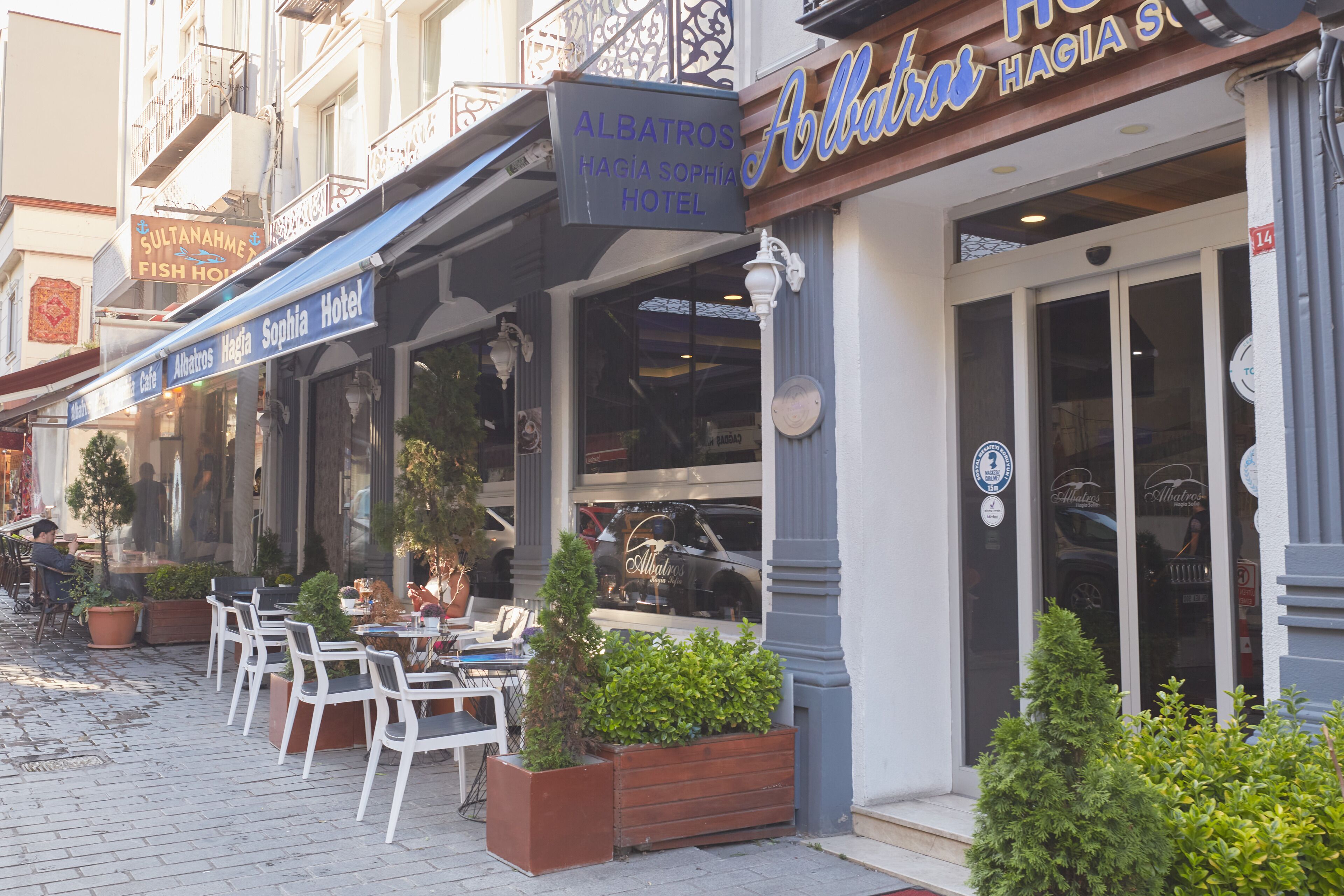 An outdoor seating area with tables and chairs, a sign for 'Albatros Hagia Sophia Hotel', and a storefront with a large glass window.