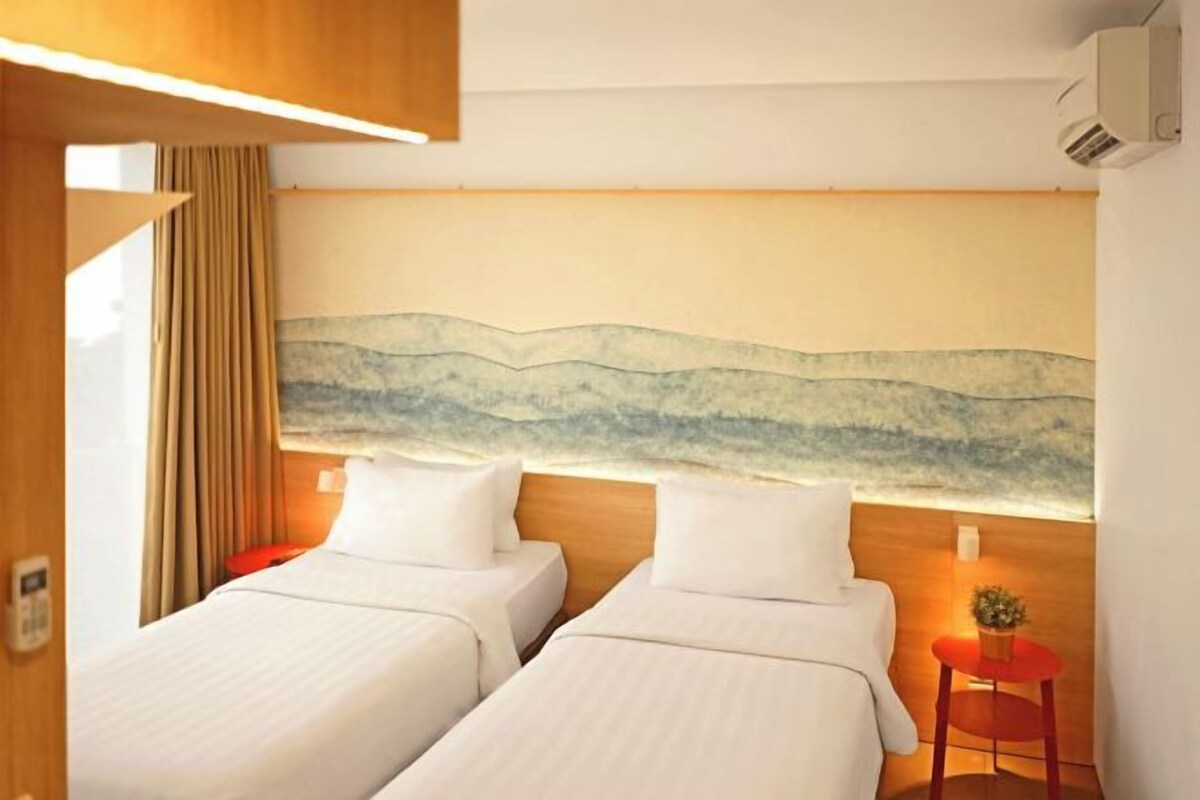Habitación de hotel con dos camas, un cabecero de madera, un cuadro colgado en la pared, una mesita con una planta y una silla roja.