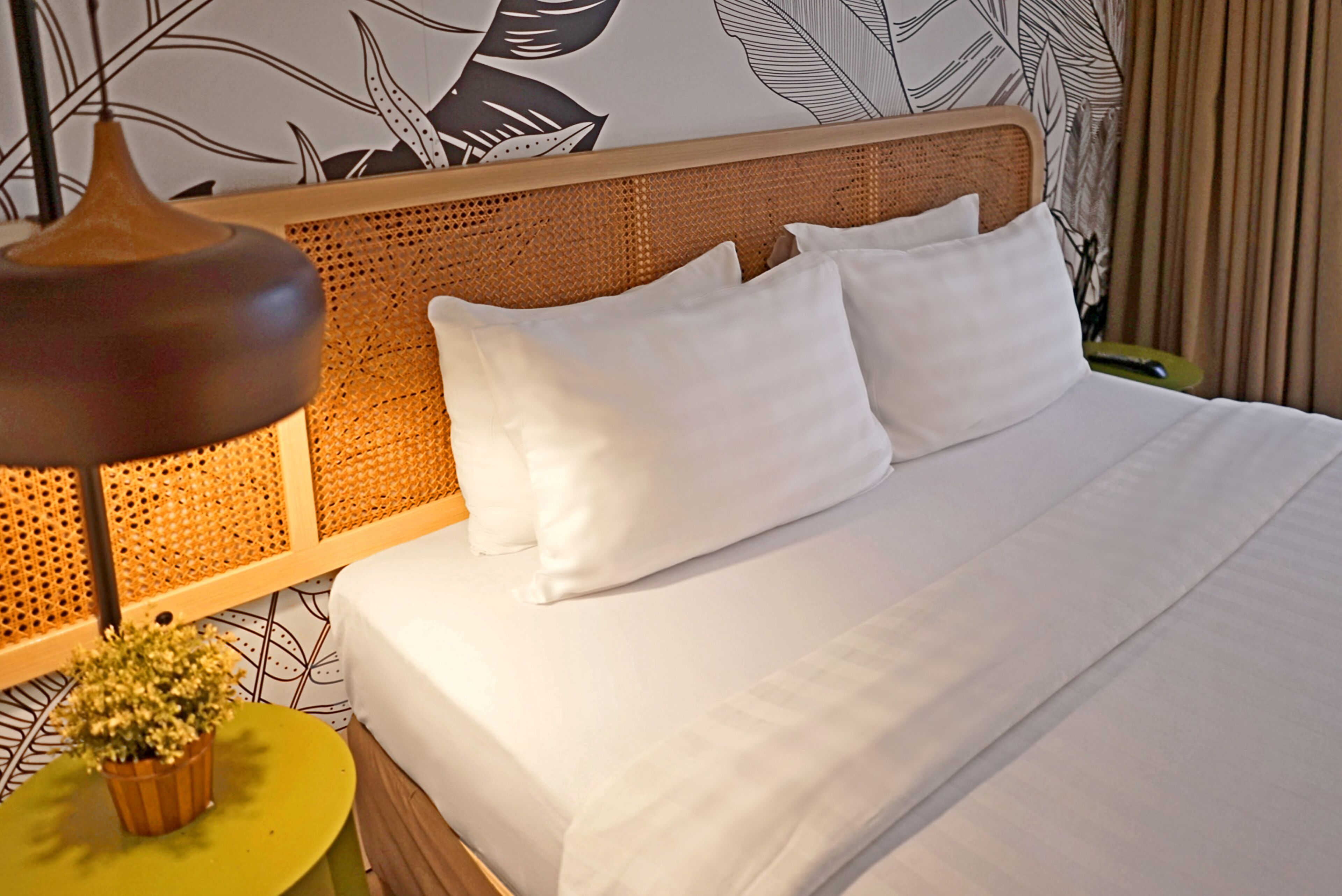 A neatly made bed with white linens and pillows, a woven headboard, a small table with a plant, and a lamp.