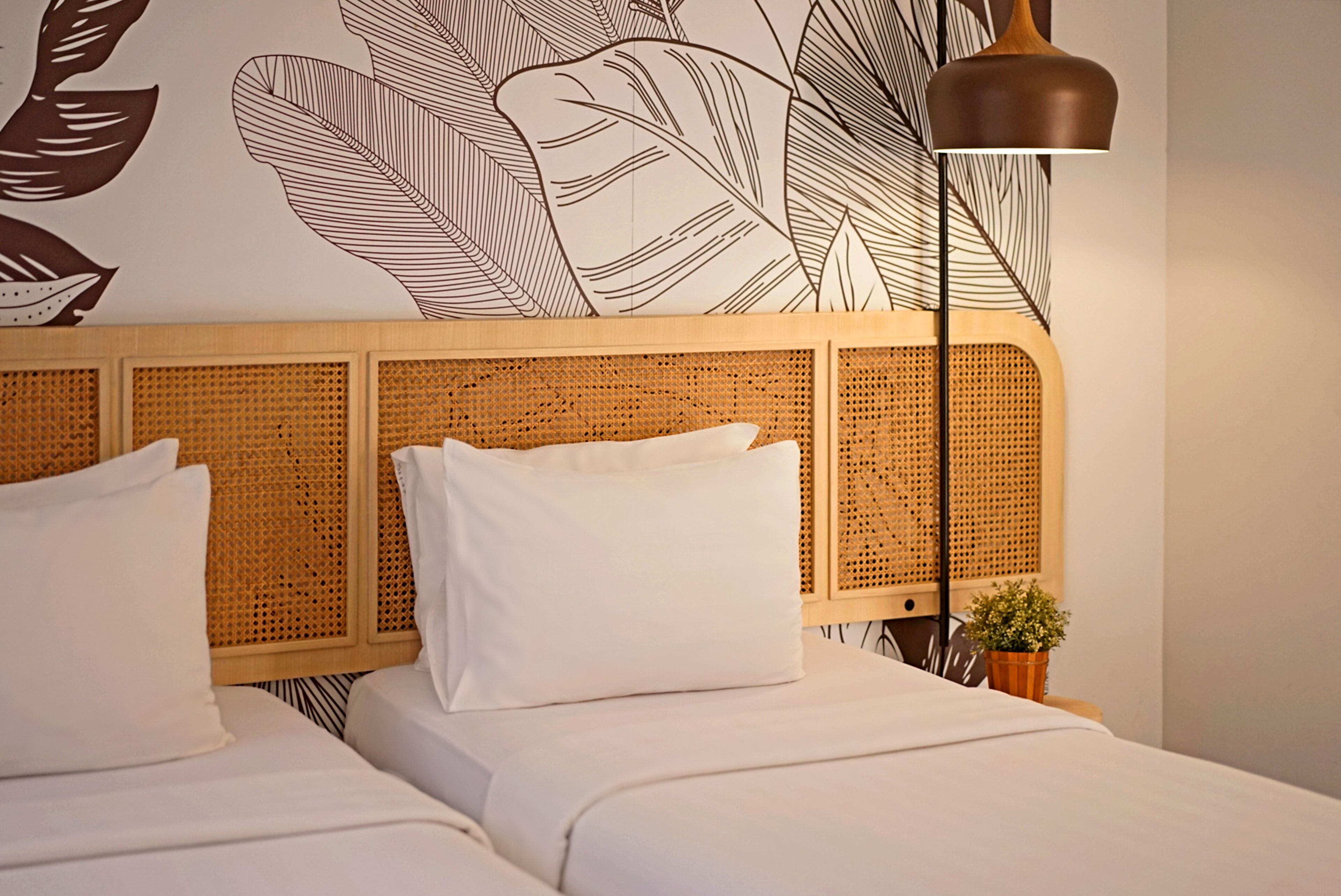 A hotel room with a woven headboard, two white beds, a small potted plant, and a floor lamp.