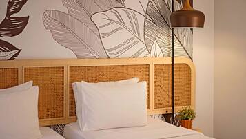 A hotel room with a woven headboard, two white beds, a small potted plant, and a floor lamp.