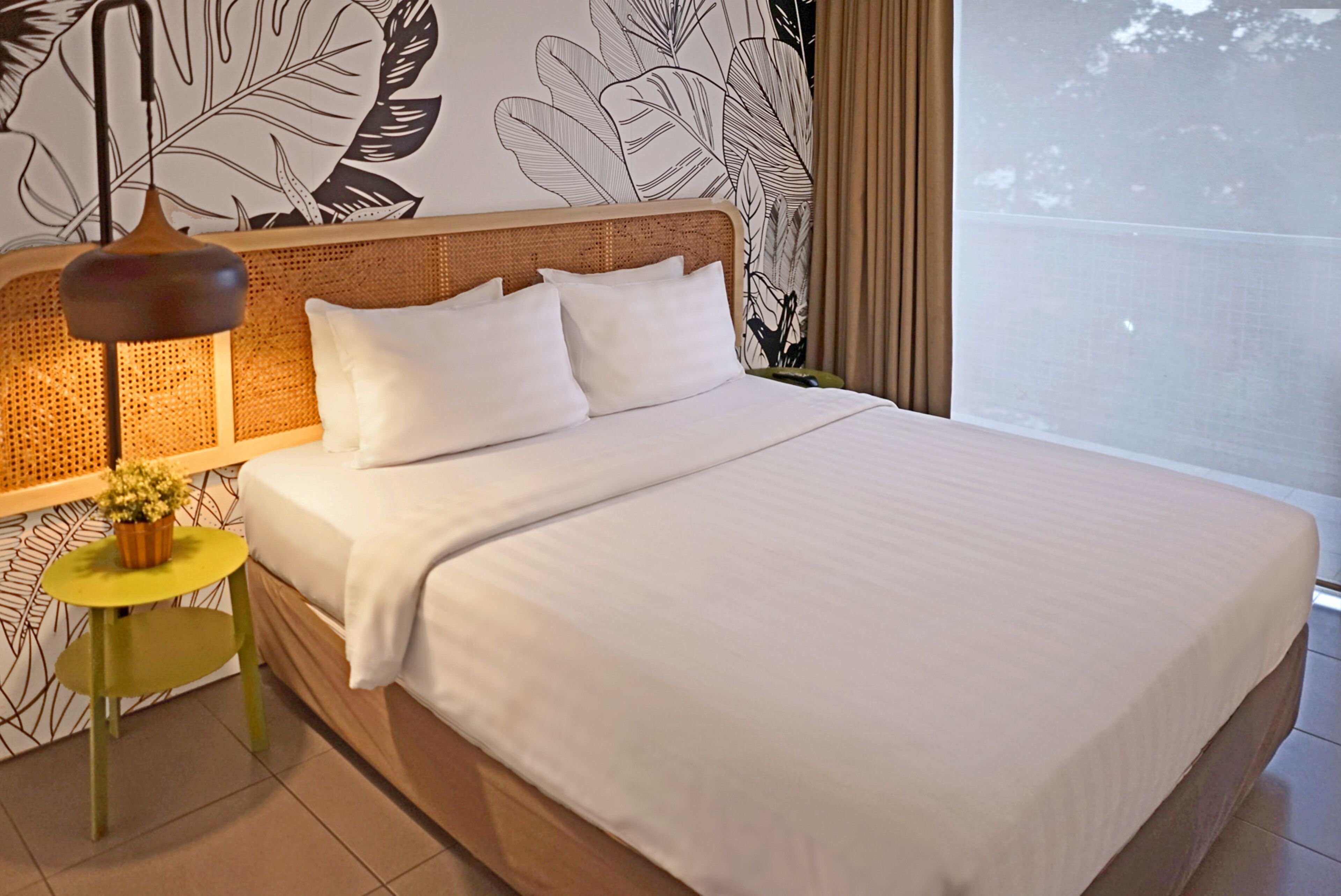 Una cama bien tendida con sábanas blancas y un cabecero de madera, una mesita redonda con una planta, y una pared con un diseño de hojas.