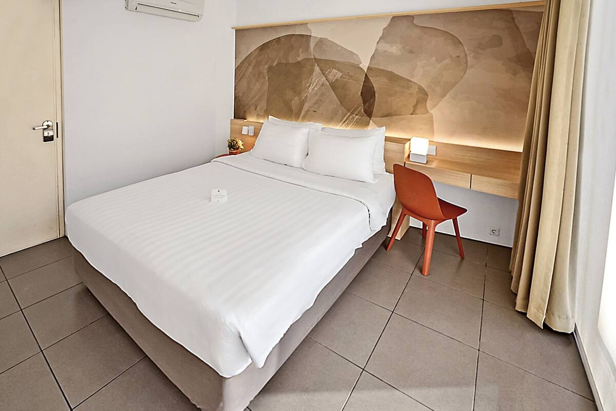 A hotel room with a large bed, a wooden headboard, a nightstand with a lamp, a chair, and a large abstract painting on the wall.