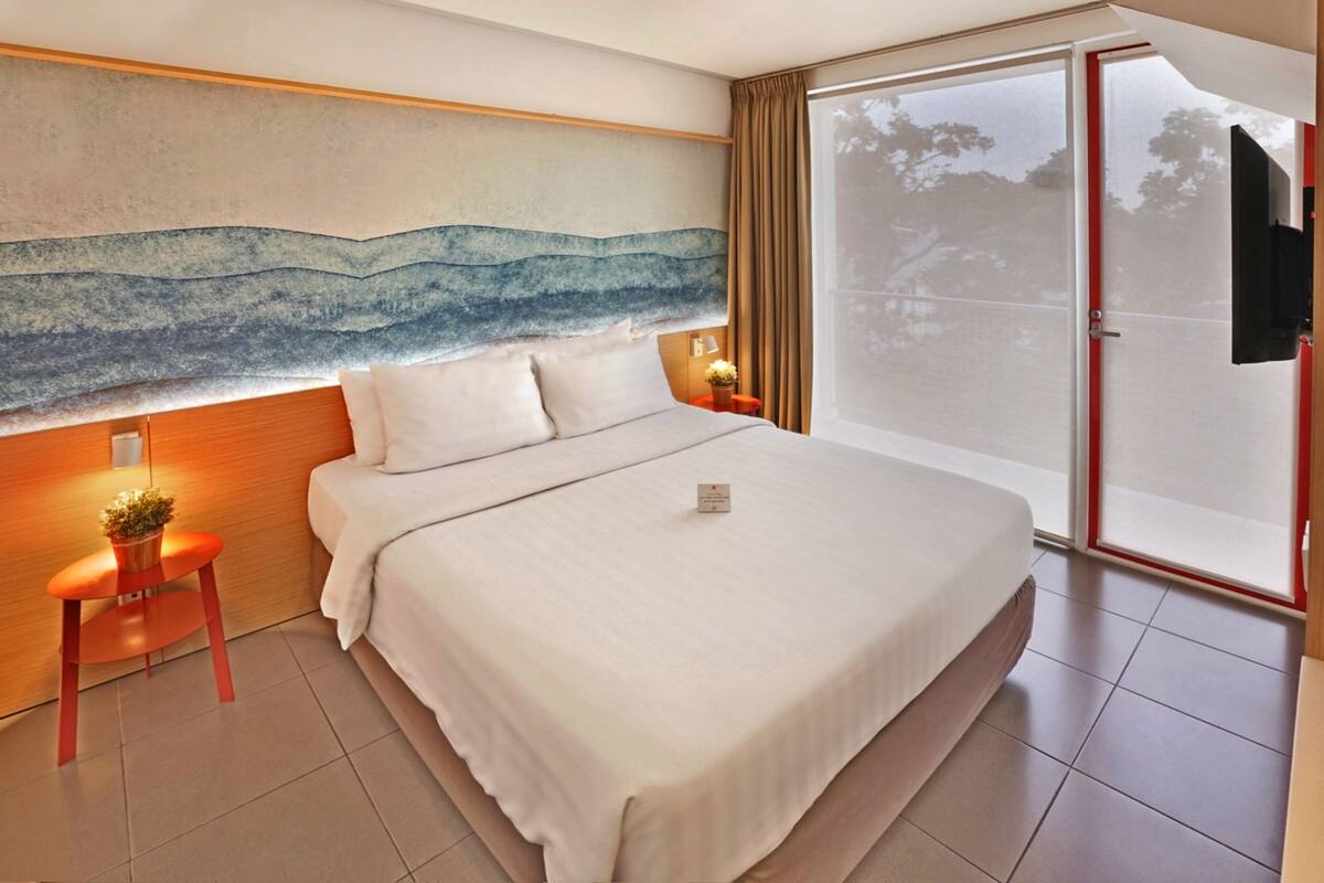A modern hotel room with a large bed, a painting on the wall, a small table with a plant, and a sliding glass door to the outside.