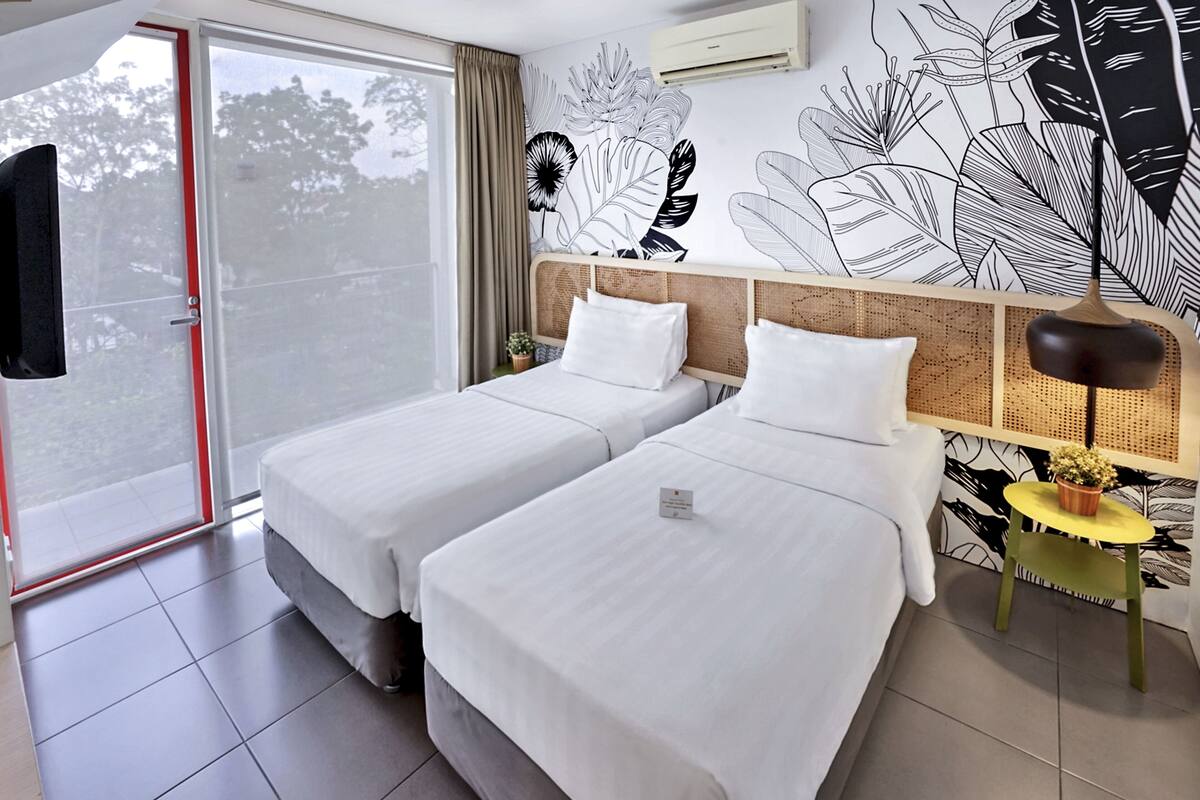 A hotel room with two beds, a large window, a wall mural, and a small table with a lamp.