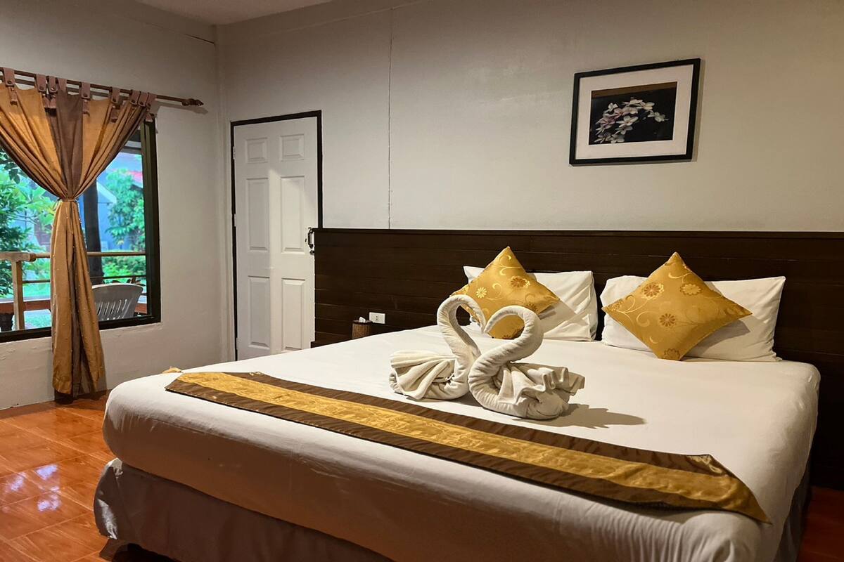Una cama bien hecha con una colcha dorada y dos almohadas doradas, un cuadro enmarcado en la pared y una ventana con cortinas.
