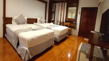 Quarto de hotel com duas camas, piso de madeira, escrivaninha com cadeira, espelho e televisão.