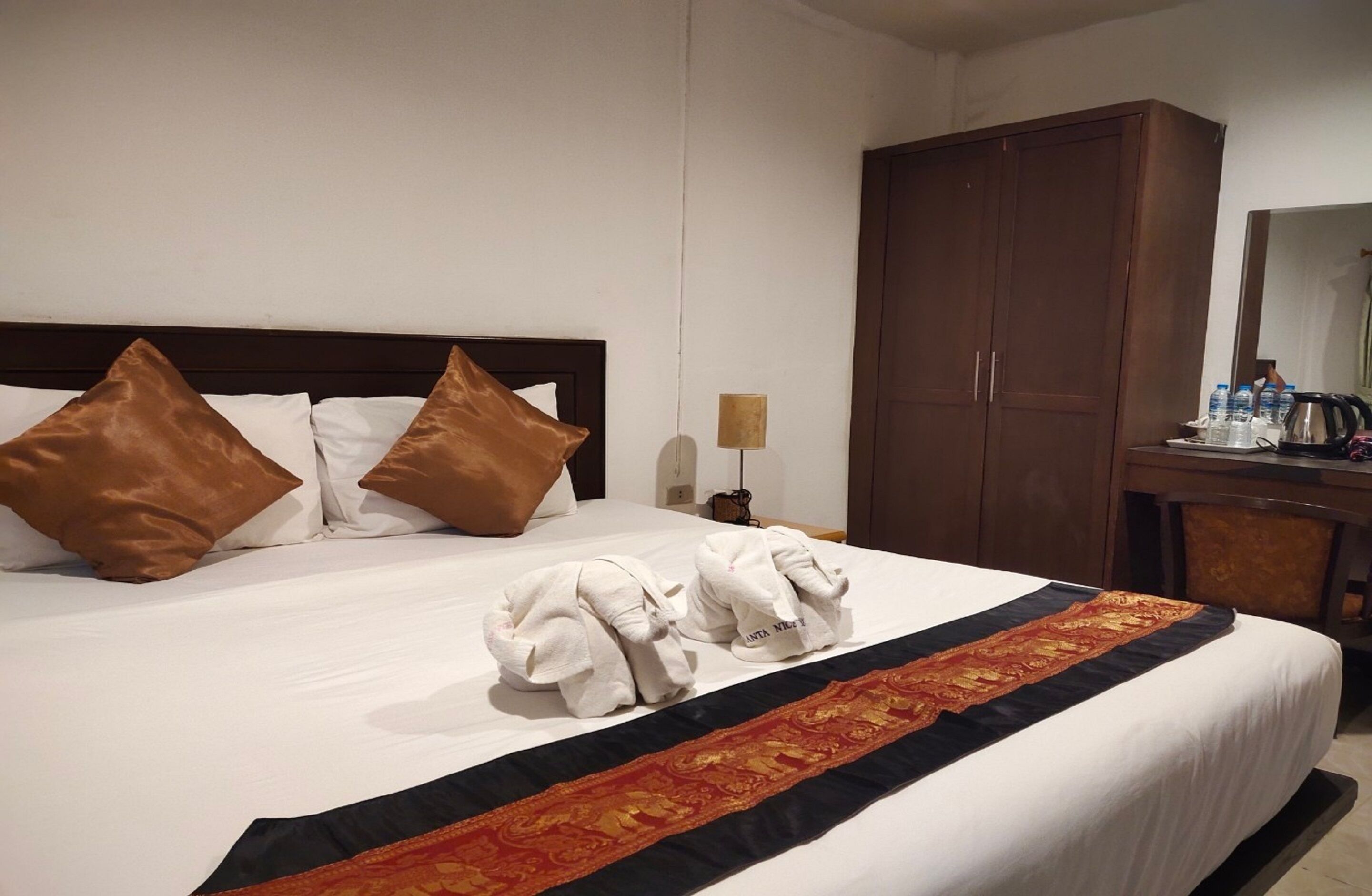A hotel room with a large bed, brown pillows, a decorative bed runner, a wooden wardrobe, and a desk with a mirror and toiletries.