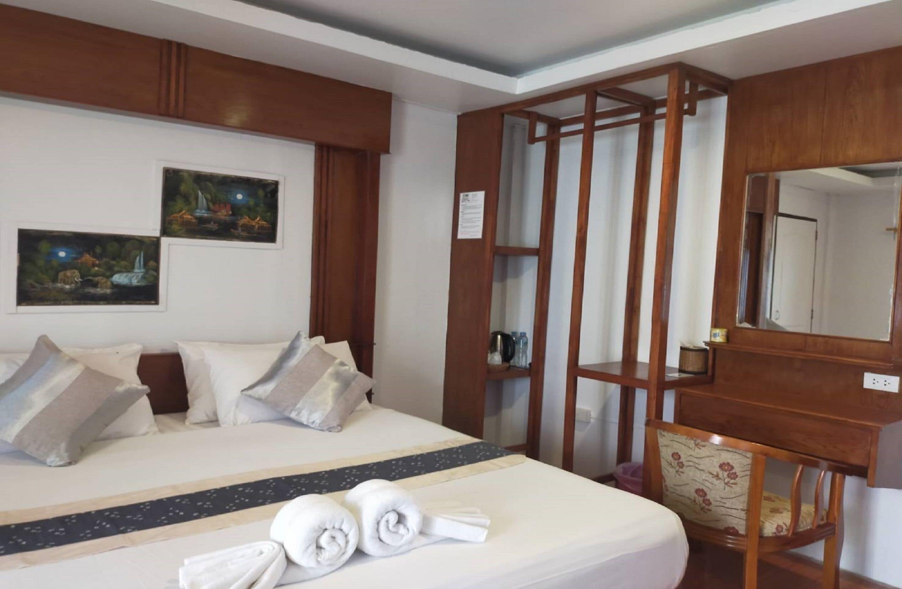 Una camera d'albergo con un letto, cuscini, un comodino in legno, uno specchio e due quadri alle pareti.