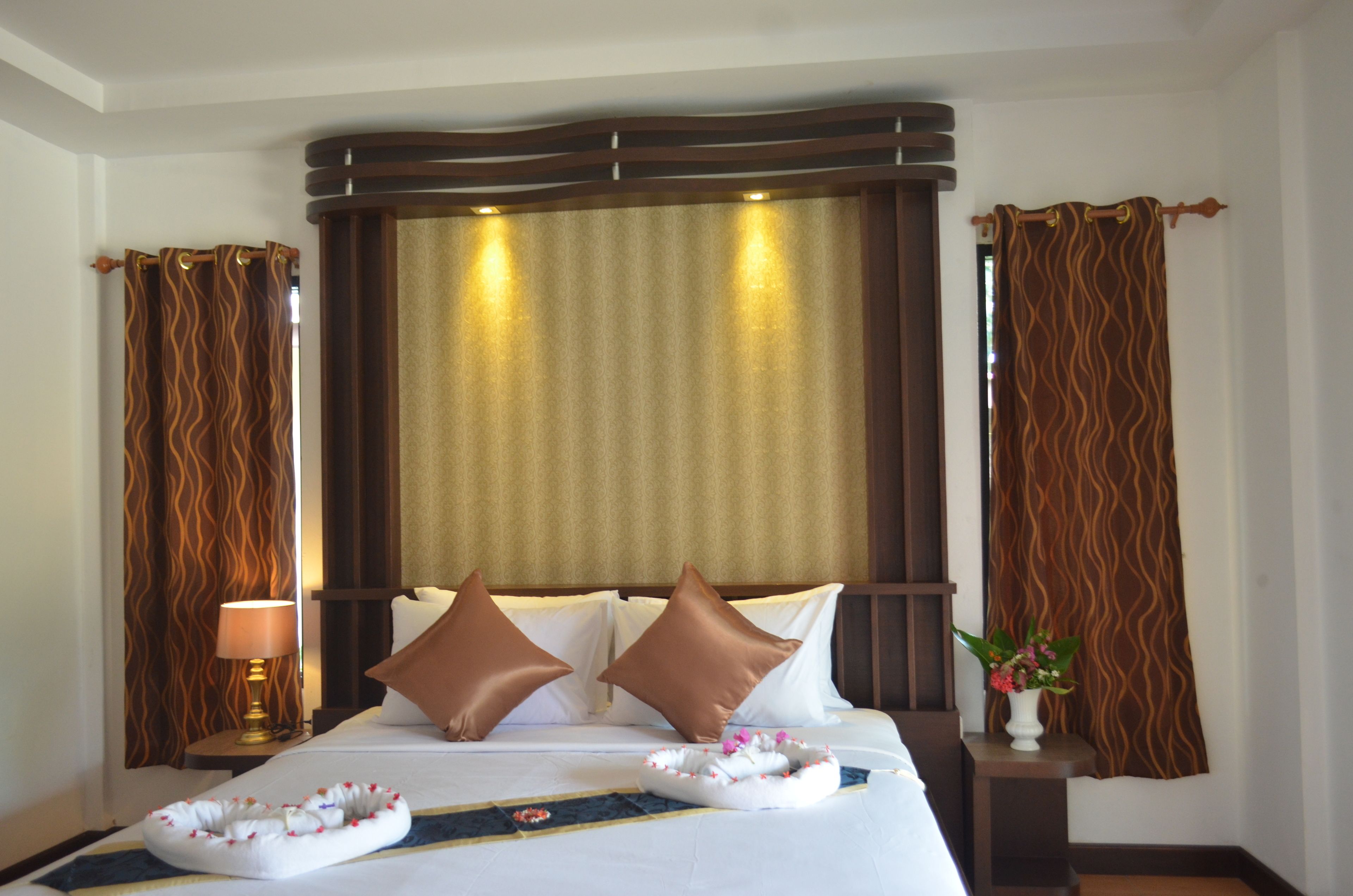 Ein Hotelzimmer mit einem großen Bett, zwei Kissen, einer Nachttischlampe, einem Zierkissen, einer Vase mit Blumen und Vorhängen.