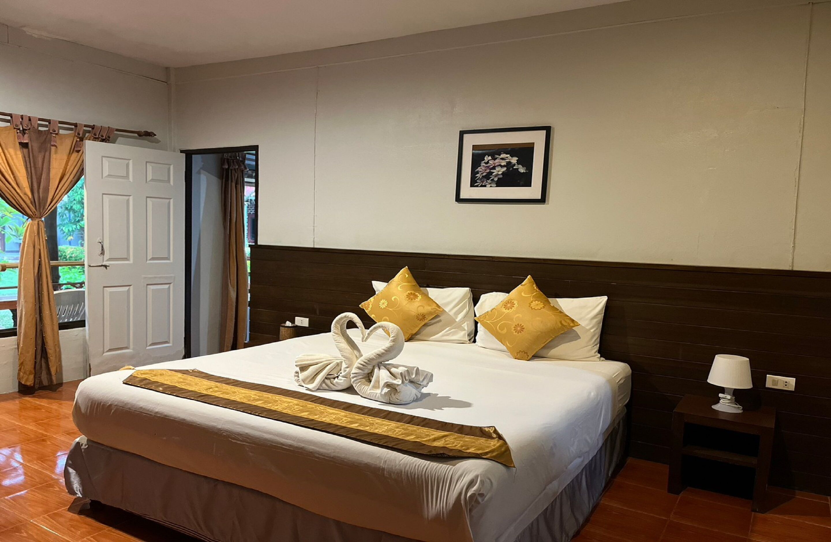 A hotel room with a large bed, a wooden headboard, a framed picture on the wall, and a nightstand with a lamp.