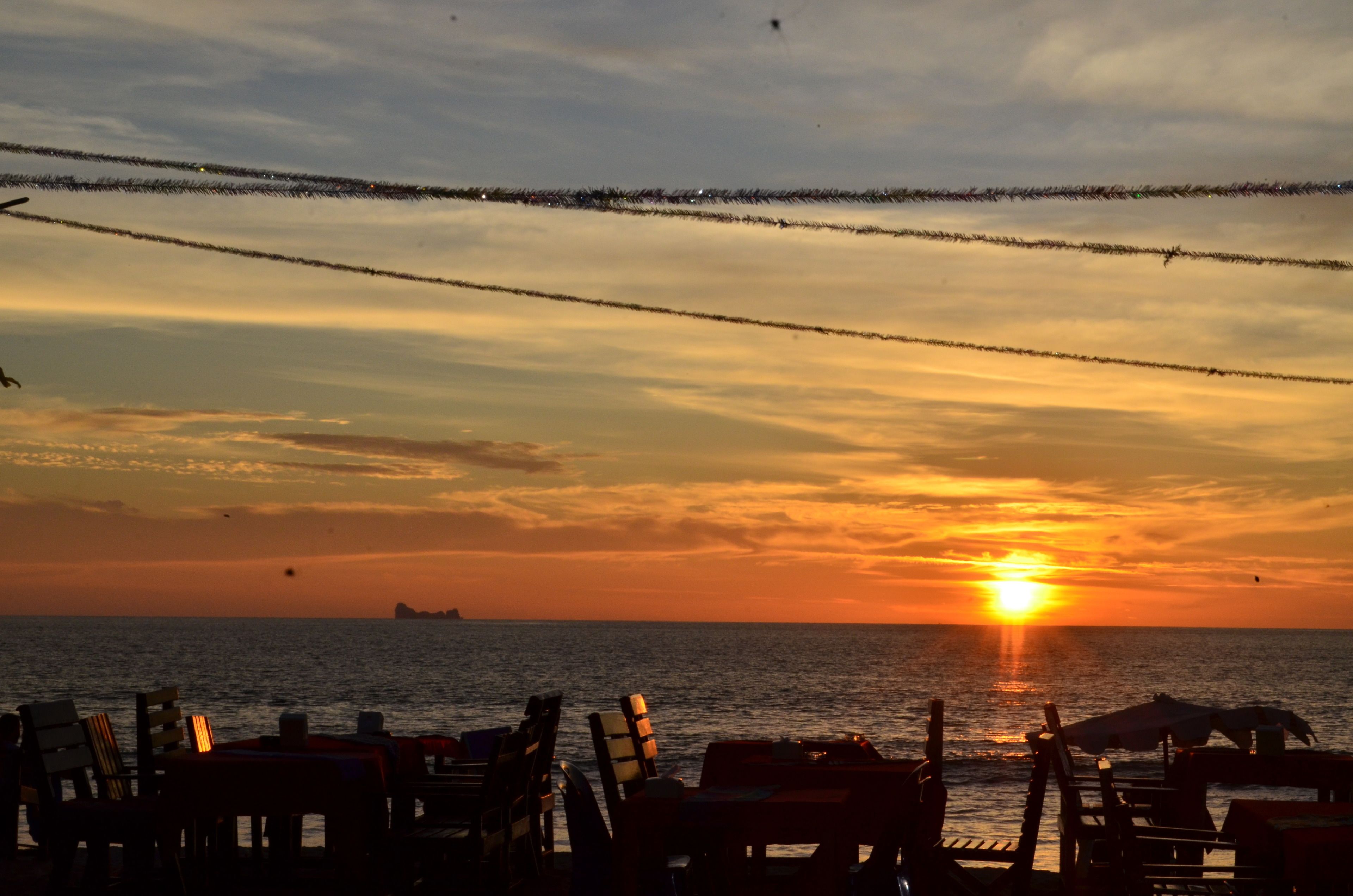 A beachside view with chairs and tables, a sunset over the ocean, and a clear sky.