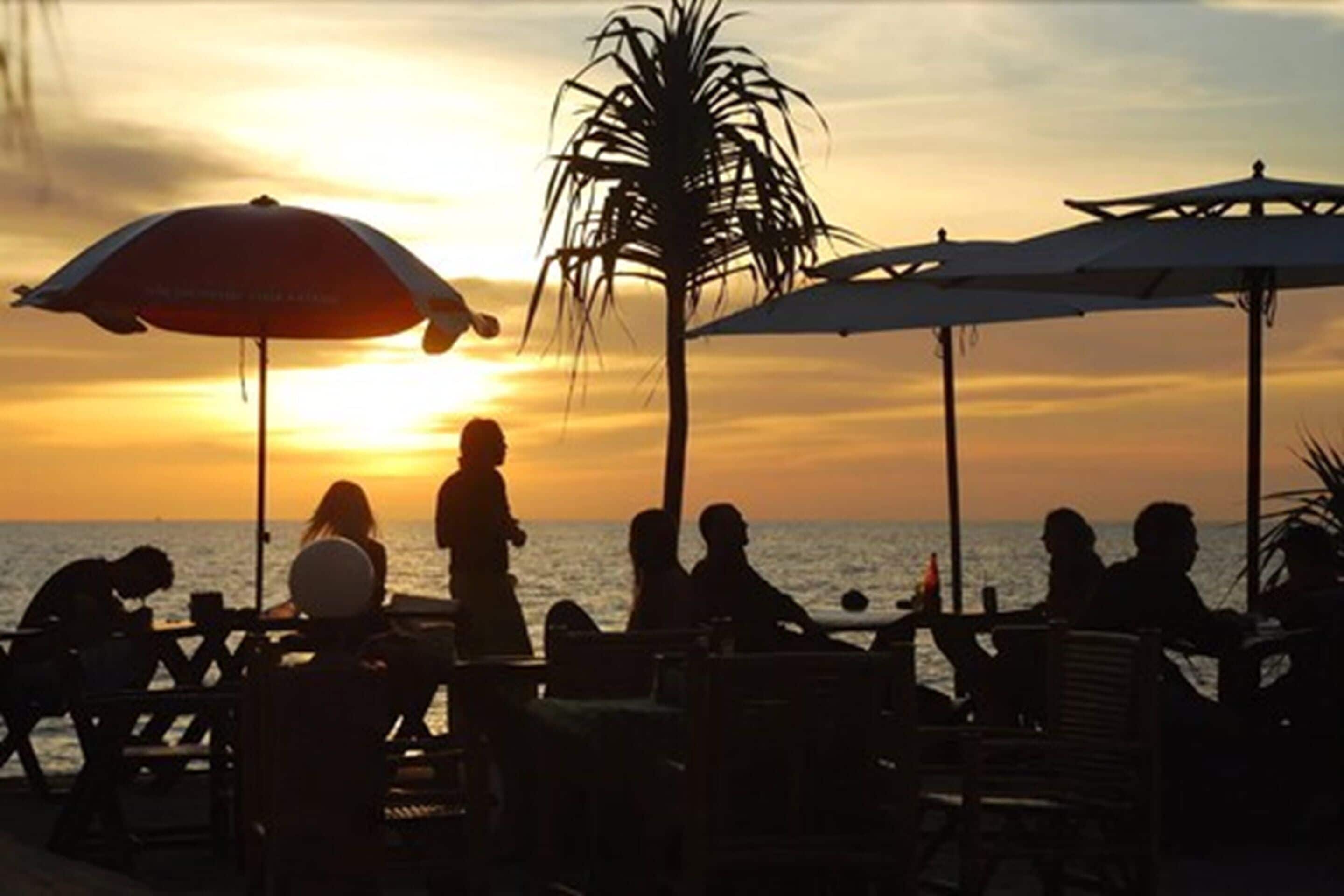 Escena en la playa con gente sentada bajo sombrillas, contemplando el océano al atardecer.
