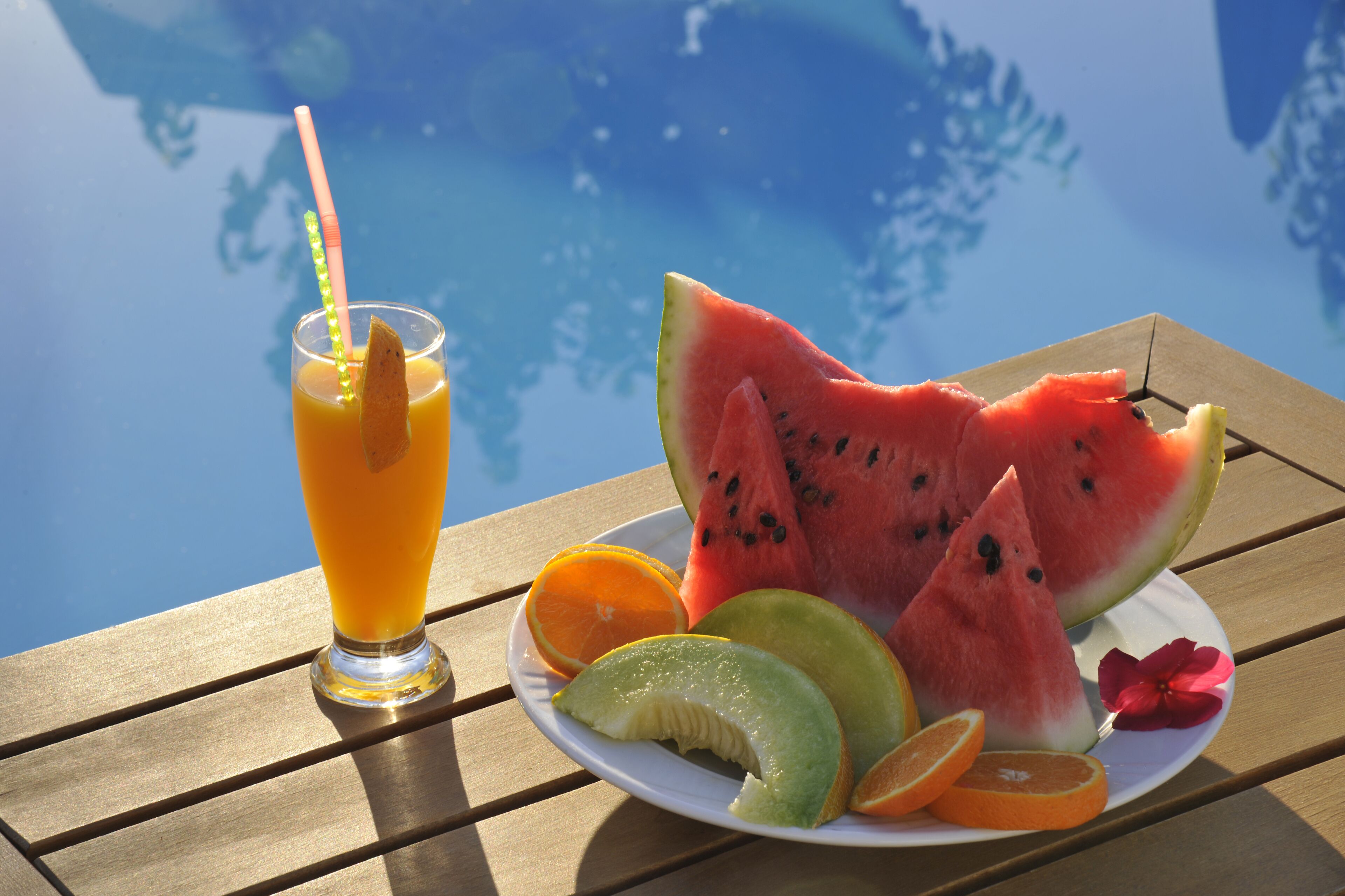 A plate of watermelon, orange, and melon slices on a wooden table by a pool.