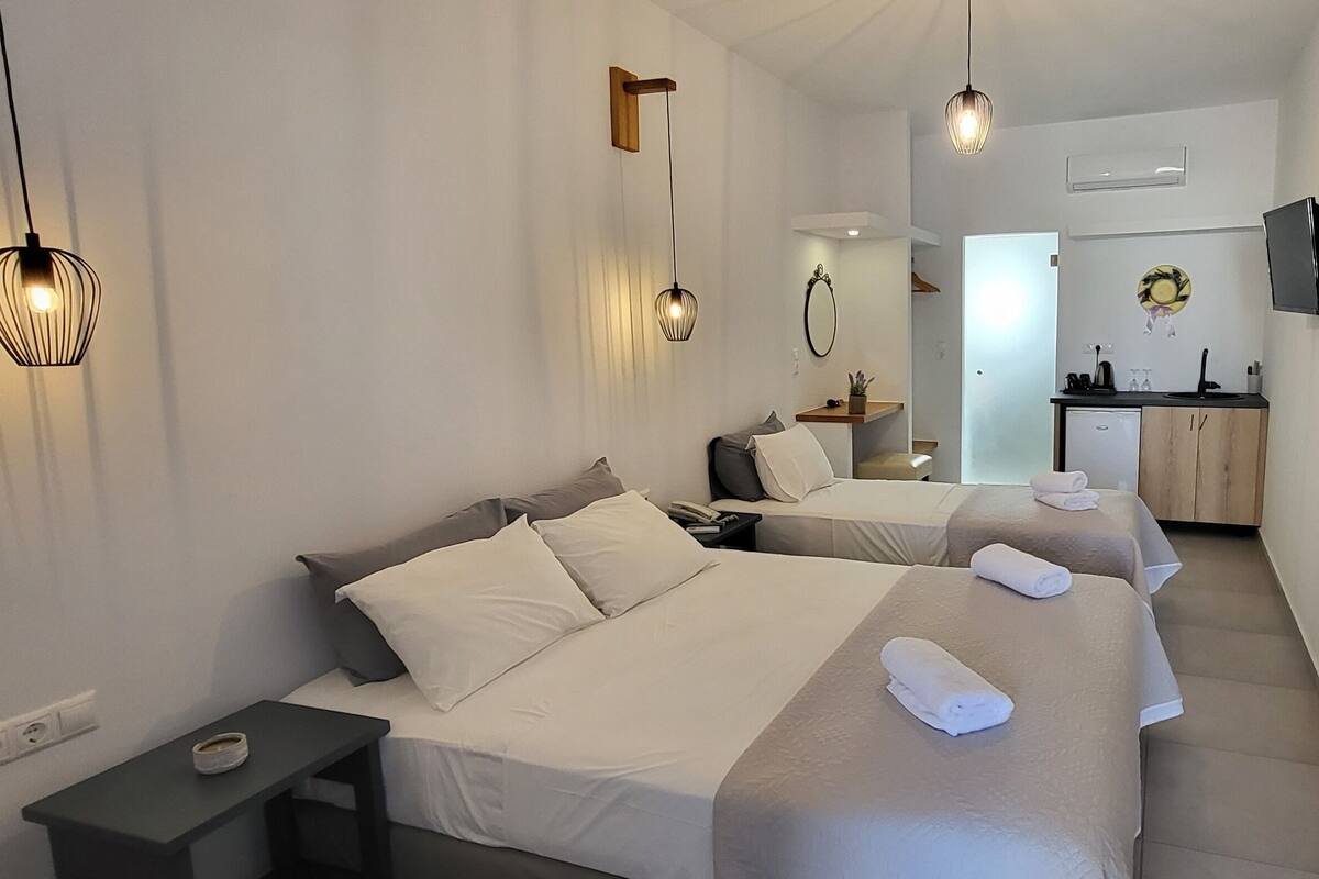 A hotel room with two beds, a small desk, a bedside table, a mirror, a TV mounted on the wall, and a kitchen area in the background.