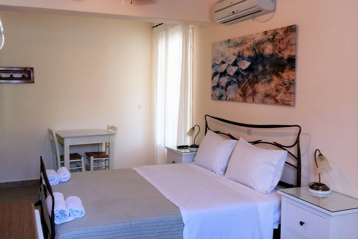 A hotel room with a bed, a bedside table, a desk, a chair, a mirror, and an air conditioning unit.