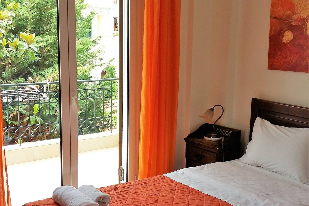 A hotel room with a bed, a nightstand, a lamp, a painting, and a view of greenery through the window.